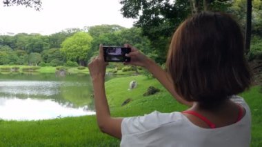 Şehir parkında akıllı telefonuyla fotoğraf çeken Asyalı bir kadın. Japon bayan dışarıda cep telefonuyla fotoğraf çekiyor. İnsanlar ve teknoloji