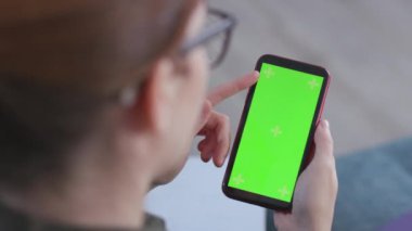 Evde akıllı telefonu olan genç, kızıl saçlı bir kadın. Yeşil ekranlı cep telefonu izleyen bir kız. Web sitesi, metin, internet, maket, e-posta, birleştirme için cep telefonu monitörünün omuz üstünden