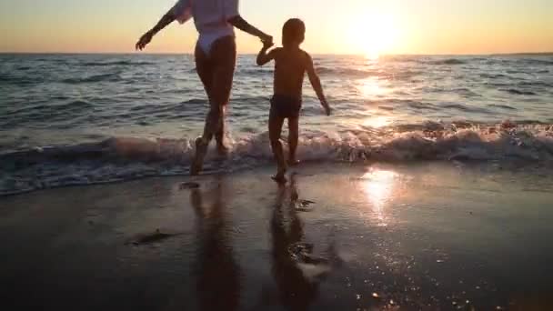 Mujer joven y su hijo vienen al agua en la puesta de sol en la playa de arena, vacaciones en la isla — Vídeos de Stock