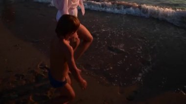 Genç bir kadın ve oğlu gün batımında kumlu sahilde, ada tatillerinde suya gelirler.