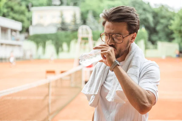 old-fashioned tennis player with towel drinking water after training on tennis court