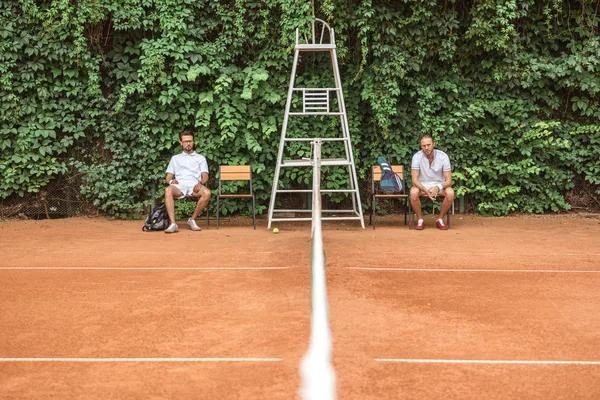 Retro styled tennis players resting after training on chairs on brown court — Stock Photo