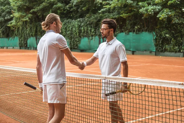 Athletic tennis players with wooden rackets shaking hands after match on court — Stock Photo
