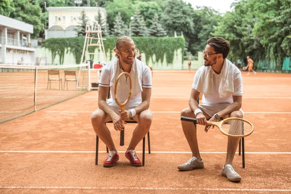 Bonitos jogadores de tênis sorridentes com raquetes de madeira descansando em cadeiras — Fotografia de Stock