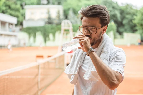 Altmodischer Tennisspieler mit Handtuch, der nach dem Training auf dem Tennisplatz Wasser trinkt — Stockfoto
