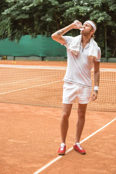 Handsome tennis player with towel drinking water on tennis court — Stock Photo