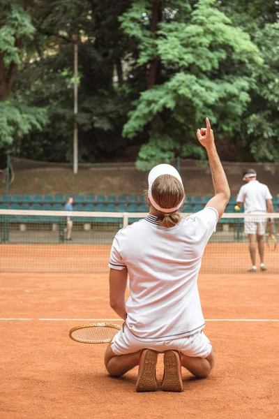 Jugador de tenis anticuado gesticulando hacia arriba y de rodillas después de ganar el partido en la cancha - foto de stock