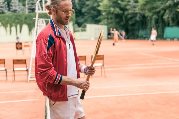 Guapo jugador de tenis a la antigua con raqueta de madera de pie en la red en la cancha de tenis - foto de stock