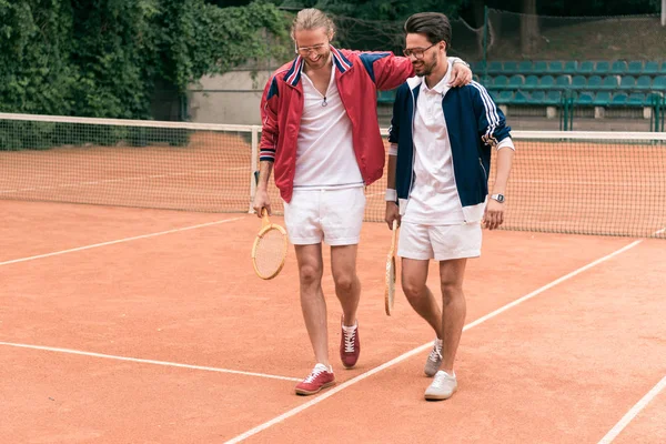 Amigos esportivos alegres com raquetes de madeira abraçando e andando no campo de ténis — Fotografia de Stock