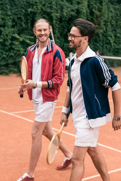 Sorridenti amici in stile retrò con racchette di legno che camminano sul campo da tennis — Foto stock