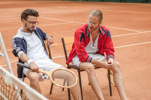 Amigos atléticos com raquetes de madeira sentados em cadeiras após o treinamento no campo de ténis — Fotografia de Stock