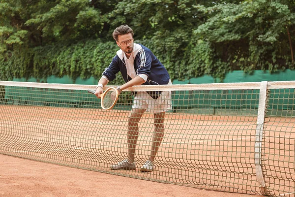 Guapo tenista con raqueta apoyada en la red de tenis en la cancha marrón - foto de stock