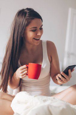 Genç kadın içme kahve evde yatağında gülümseyen ve onun smartphone kontrol