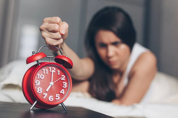 Angry young woman kill off alarm clock by fist on the bed in the morning. Awakening of the sleeping woman.