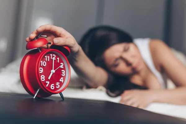 Angry young woman switches off the alarm clock on the bed in the morning. Awakening of the sleeping woman. Focus on alarm clock.