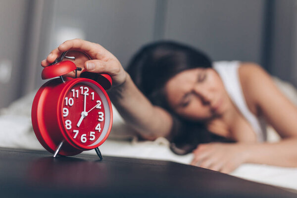 Angry young woman switches off the alarm clock on the bed in the morning. Awakening of the sleeping woman. Focus on alarm clock.