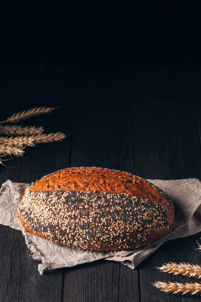 Laib braunes Brot mit Sesam und Mohn auf Textiltuch auf schwarzem Holzhintergrund mit Kopierraum — Stockfoto