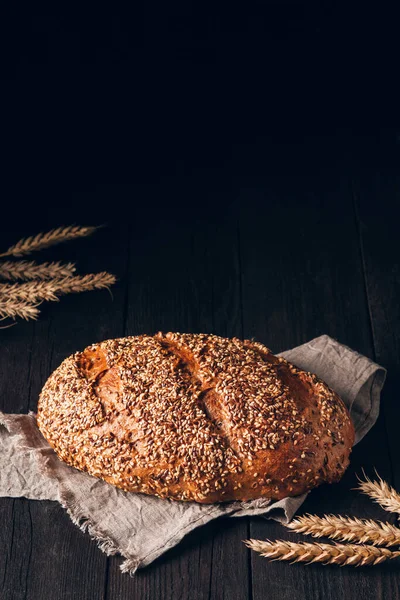 Laib braunes Brot mit Sesam und Leinsamen auf Textiltuch auf schwarzem Holzhintergrund mit Kopierraum lizenzfreie Stockfotos