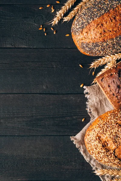 Panes negros y marrones con espiguillas sobre fondo de madera con espacio para copiar. Tres tipos diferentes de pan a la derecha. Panadería, cocina y concepto de tienda de comestibles —  Fotos de Stock