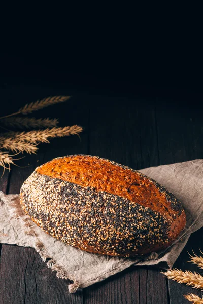Laib braunes Brot mit Sesam und Mohn auf Textiltuch auf schwarzem Holzhintergrund mit Kopierraum — Stockfoto