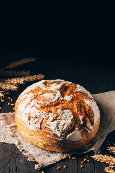 Laib braunes Brot mit Rissen und Mehl auf Textiltuch auf schwarzem Holzhintergrund — Stockfoto