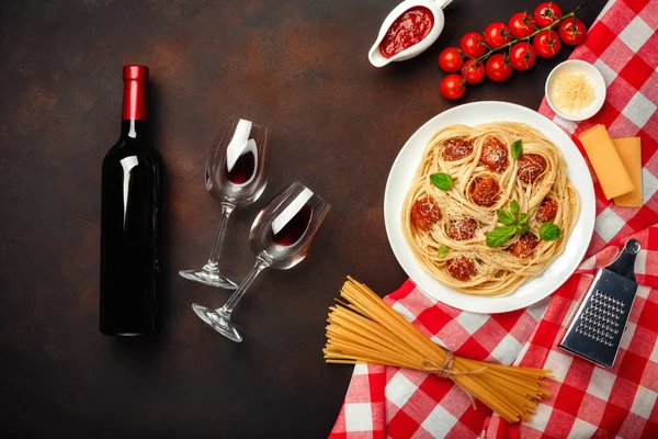 Spaghetti pasta with meatballs, cherry tomato sauce, cheese, wineglass and bottle on rusty background, top view.