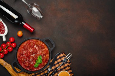 Baharatlar, cherry domates, makarna ve fesleğen ile bir şişe şarap ve bardağı paslı kahverengi zemin üzerinde bir tavada domates soslu köfte. Üstten görünüm.