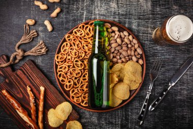 Bir şişe bira bir plaka ile tuzlu ookies simit, fıstık fındık ve cips siyah çizik tebeşir tahtası üzerinde. Üstten görünüm.