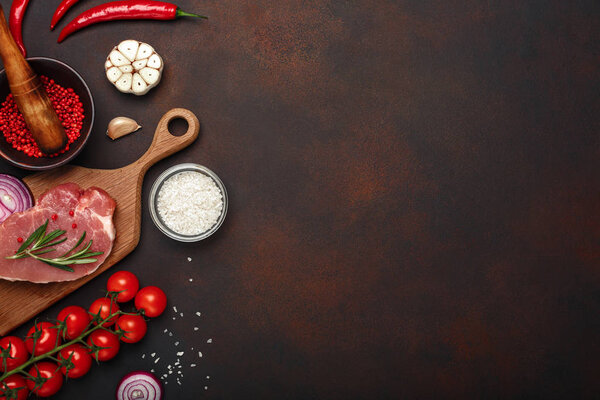 Pieces of raw pork steak on cutting board with cherry tomatoes, rosemary, garlic, red pepper, onion, salt and spice mortar on rusty brown background. Top view with space for your text.