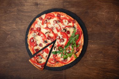 Pizza Tütsülenmiş sosis, peynir, mantar, cherry domates, biber ve yeşillik bir taş ve ahşap kahverengi masa, üstten görünüm.