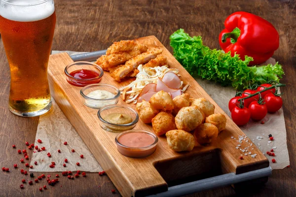 Big set of snacks for beer it includes fried cheese balls, pigtail cheese, ham and crab sticks served with garlic sauces, ketchup and mustard on a wooden board and brown background.