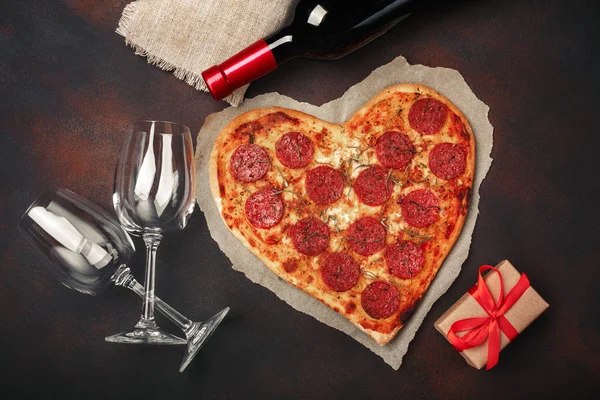 Heart shaped pizza with mozzarella, sausagered, wine bottle, two wineglass, gift box on rusty background. Top view.