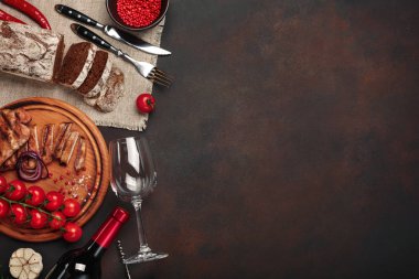 Izgara domuz biftek şişe şarap, şarap kadehi, tirbuşon, bıçak, çatal, siyah ekmek, domates, sarımsak, soğan ve biberiye ile paslı arka plan üzerine dilimlenmiş. Üstten görünüm.