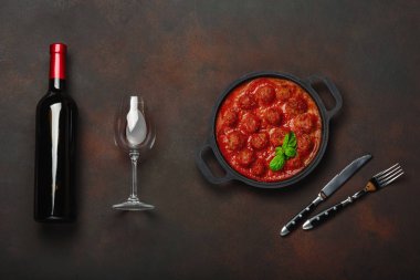 Baharatlar, cherry domates, makarna ve fesleğen ile bir şişe şarap ve bardağı paslı kahverengi zemin üzerinde bir tavada domates soslu köfte. Üstten görünüm.