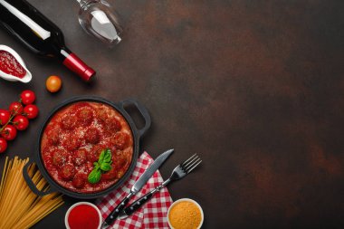 Baharatlar, cherry domates, makarna ve fesleğen ile bir şişe şarap ve bardağı paslı kahverengi zemin üzerinde bir tavada domates soslu köfte. Üstten görünüm.