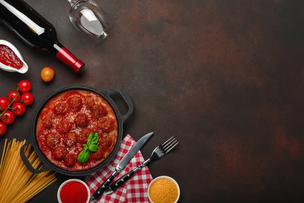 Baharatlar, cherry domates, makarna ve fesleğen ile bir şişe şarap ve bardağı paslı kahverengi zemin üzerinde bir tavada domates soslu köfte. Üstten görünüm.