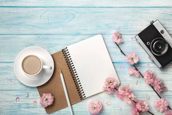 Sakura çiçekler, defter ve mavi ahşap bir masa üzerinde eski fotoğraf makinesi ile beyaz fincan kapuçino. Metin alanı olan üstten görünüm.