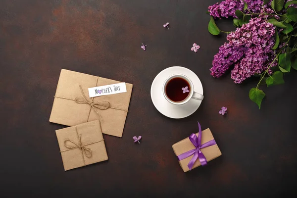 A bouquet of lilacs with cup of tea, gift box, craft envelope, a love note on rusty background. Top view with place for your text. Mother\'s day.