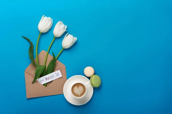 Beyaz Lale yaprakları, kahve, macarons, bir aşk notu ve zarfın üzerinde mavi bir arka plan ile. Üstten görünüm kutlama için yer ile. Anneler günü.