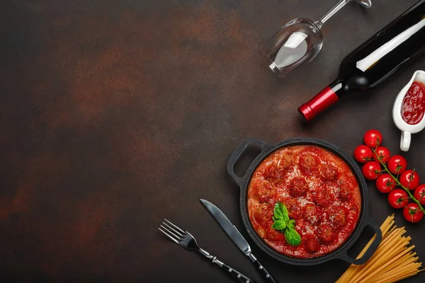 Meatballs in tomato sauce with spices, cherry tomatoes, pasta and basil in a frying pan with bottle of wine and wineglass on rusty brown background. Top view.