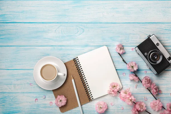 Sakura çiçekler, defter ve mavi ahşap bir masa üzerinde eski fotoğraf makinesi ile beyaz fincan kapuçino. Metin alanı olan üstten görünüm.