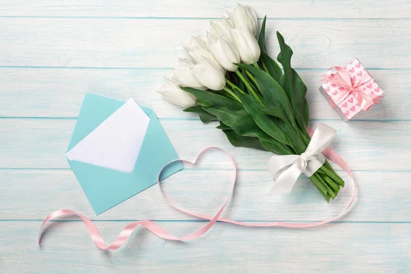 A bouquet of white tulips and a pink ribbon in the form of a heart with a gift box, love note and color envelope on blue wooden boards — Stock Photo, Image
