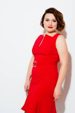 Charming plus size model in beautiful red dress looking at camera while standing on white background clipart