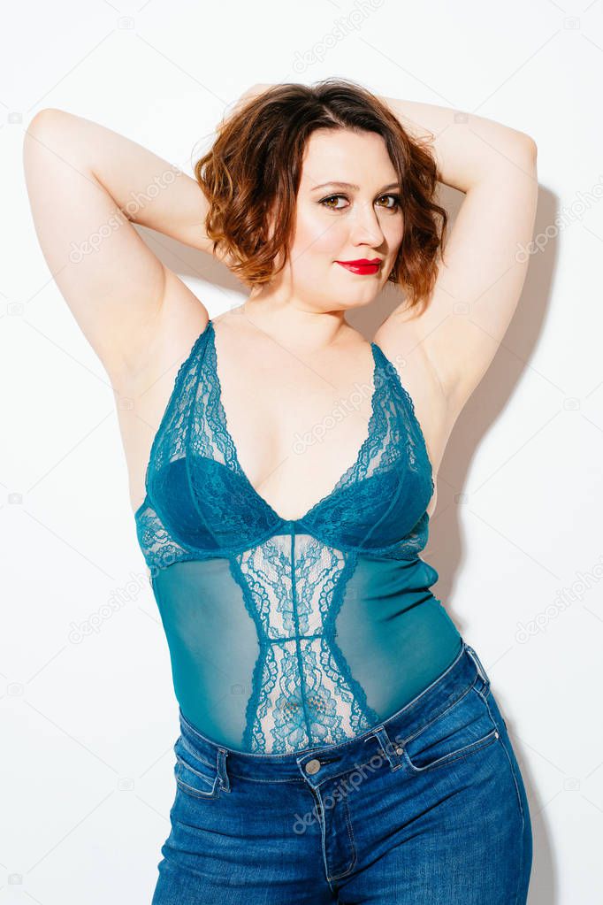 Lovely plump woman in lace lingerie and jeans keeping hands behind head and looking at camera while standing on white background