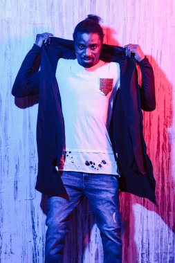 Afrikalı-Amerikalı erkek renkli neon aydınlatma duvara yakın duran ve kameraya seyir siyah hoodie koyarak ürün görünümü