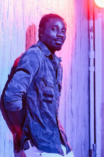 カラフルなネオン照明とカメラ目線で壁にもたれてイヤリング立っているアフリカ系アメリカ人男性 — ストック写真