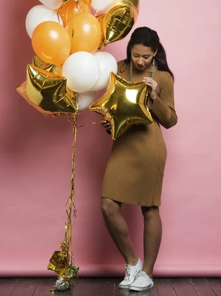 Cute young ethnicfemale in trendy knitted dress and white sneakers looking at bright shiny golden star shaped balloon in hand hanging in bunch on pink background