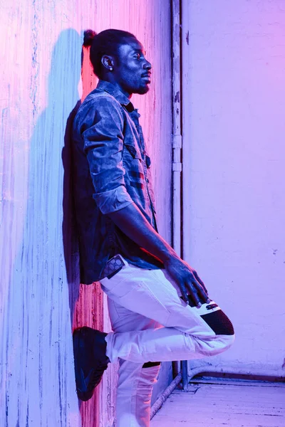 Side view of African-American man leaning on wall with leg up standing on neon illumination