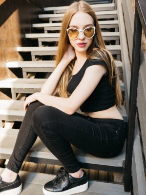 Siyah aşınma ve güneş gözlüğü merdivenlerde oturan uzun sarı saçlı trendy rahat genç kadın portresi