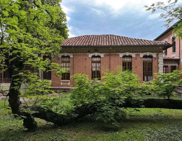 Traditional colonial house made of bricks and roof of clay tiles. Latin and Spanish colonial architecture. Garden with tree in front of the old house.